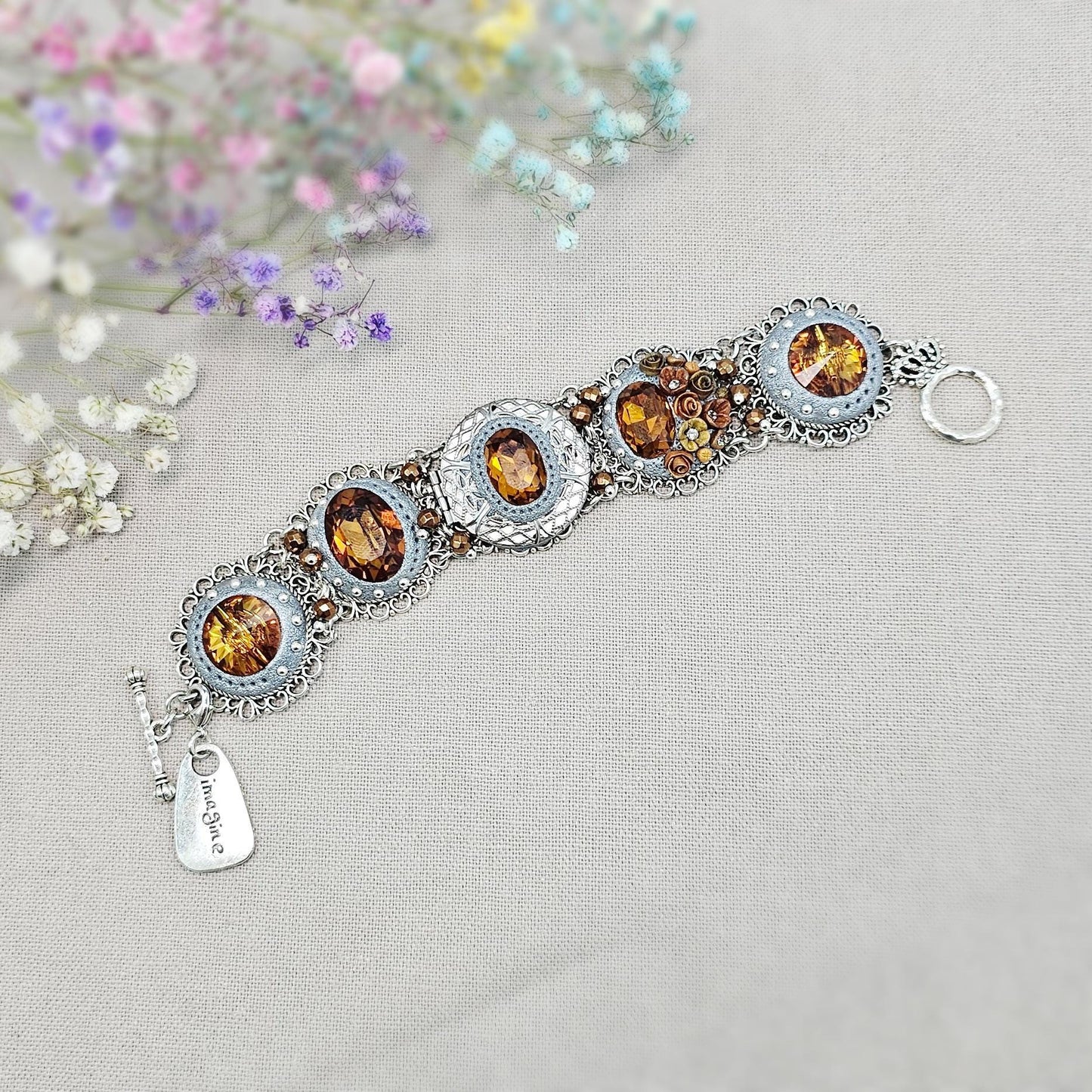 Amber-tone wide bracelet with sparkling crystals and intricate details. Features handmade polymer clay flowers, a functional locket with a red heart inside, and a detachable "imagine" charm.