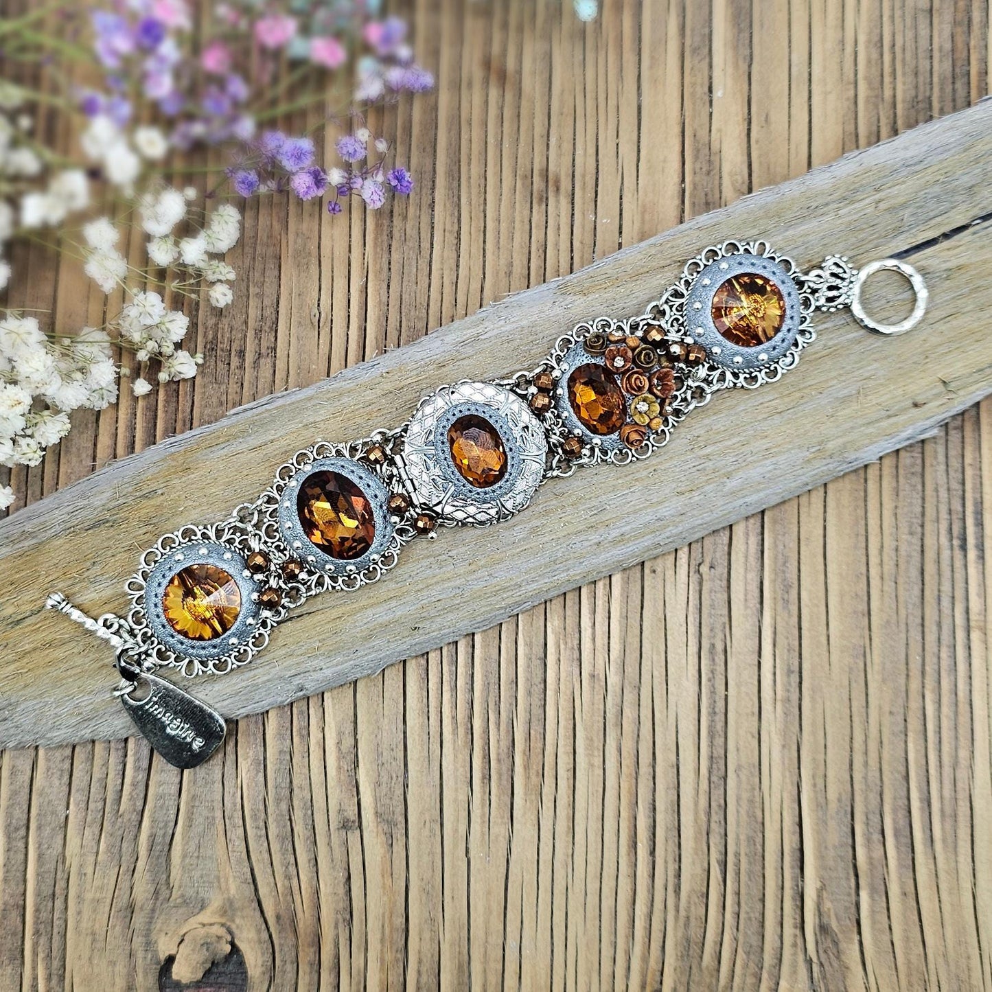 Amber-tone wide bracelet with sparkling crystals and intricate details. Features handmade polymer clay flowers, a functional locket with a red heart inside, and a detachable "imagine" charm.