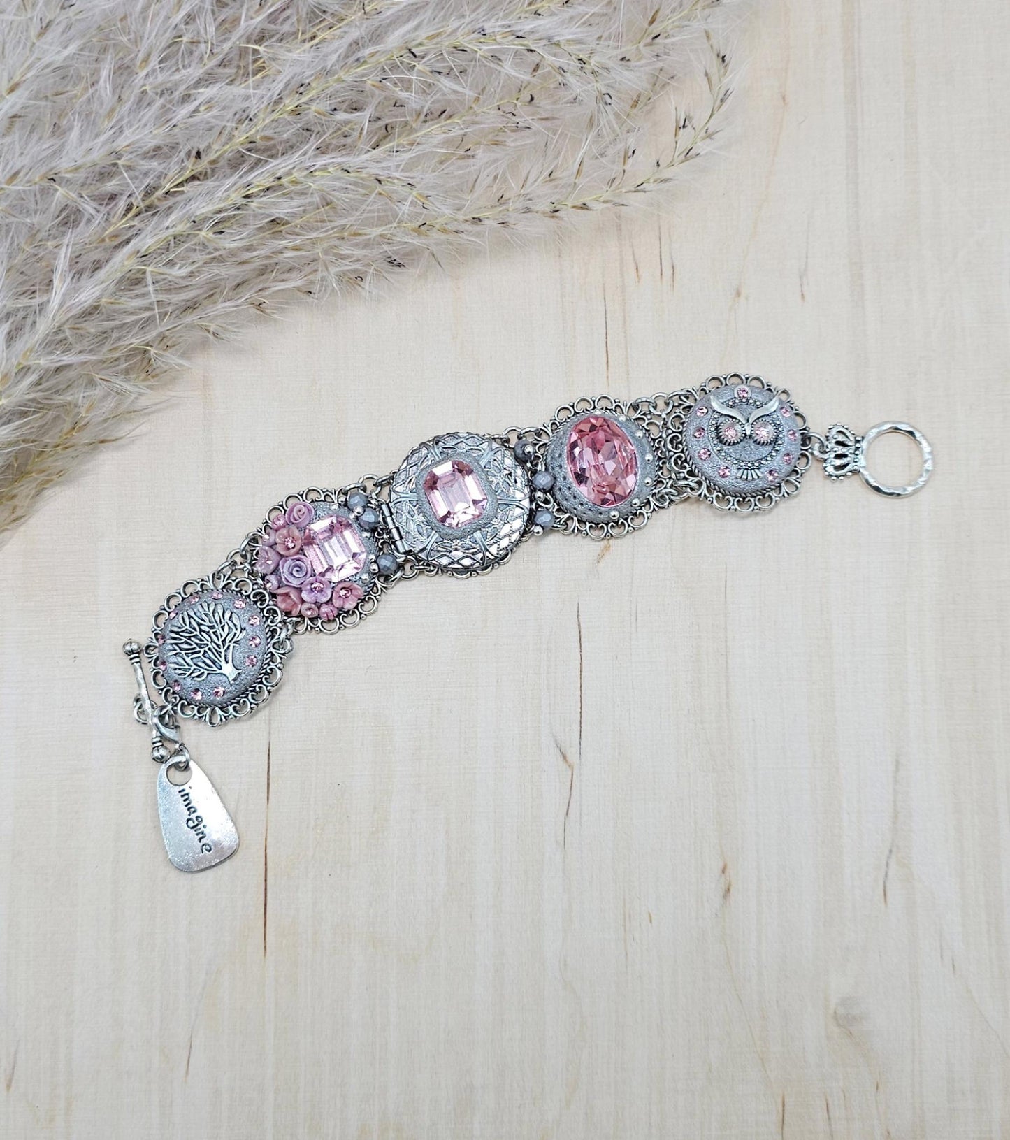 A handmade wide bracelet with pink crystals, polymer clay flowers, a Tree of Life charm, and an owl charm. Features a locket centerpiece with a hidden heart and a removable "Imagine" tag. Elegant and symbolic, perfect as a meaningful gift.