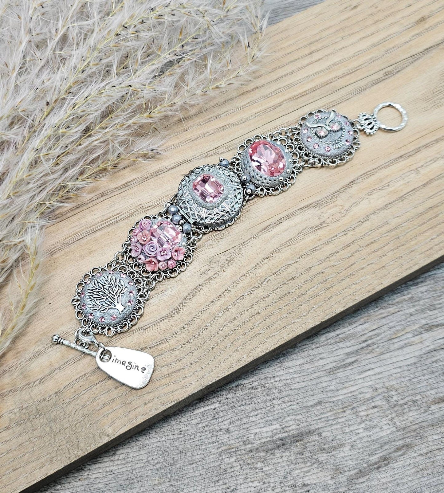 A handmade wide bracelet with pink crystals, polymer clay flowers, a Tree of Life charm, and an owl charm. Features a locket centerpiece with a hidden heart and a removable "Imagine" tag. Elegant and symbolic, perfect as a meaningful gift.