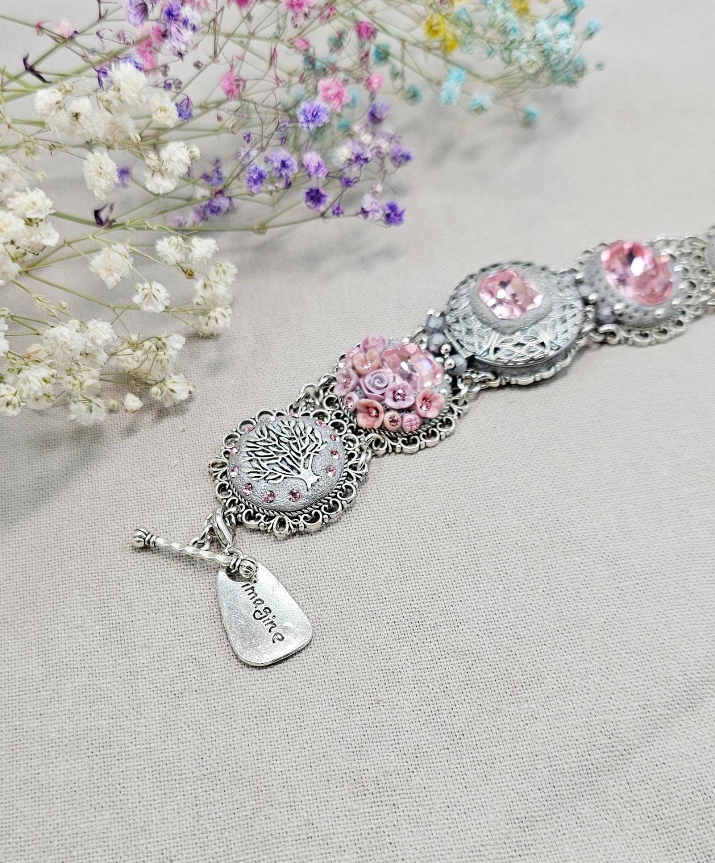 A handmade wide bracelet with pink crystals, polymer clay flowers, a Tree of Life charm, and an owl charm. Features a locket centerpiece with a hidden heart and a removable "Imagine" tag. Elegant and symbolic, perfect as a meaningful gift.