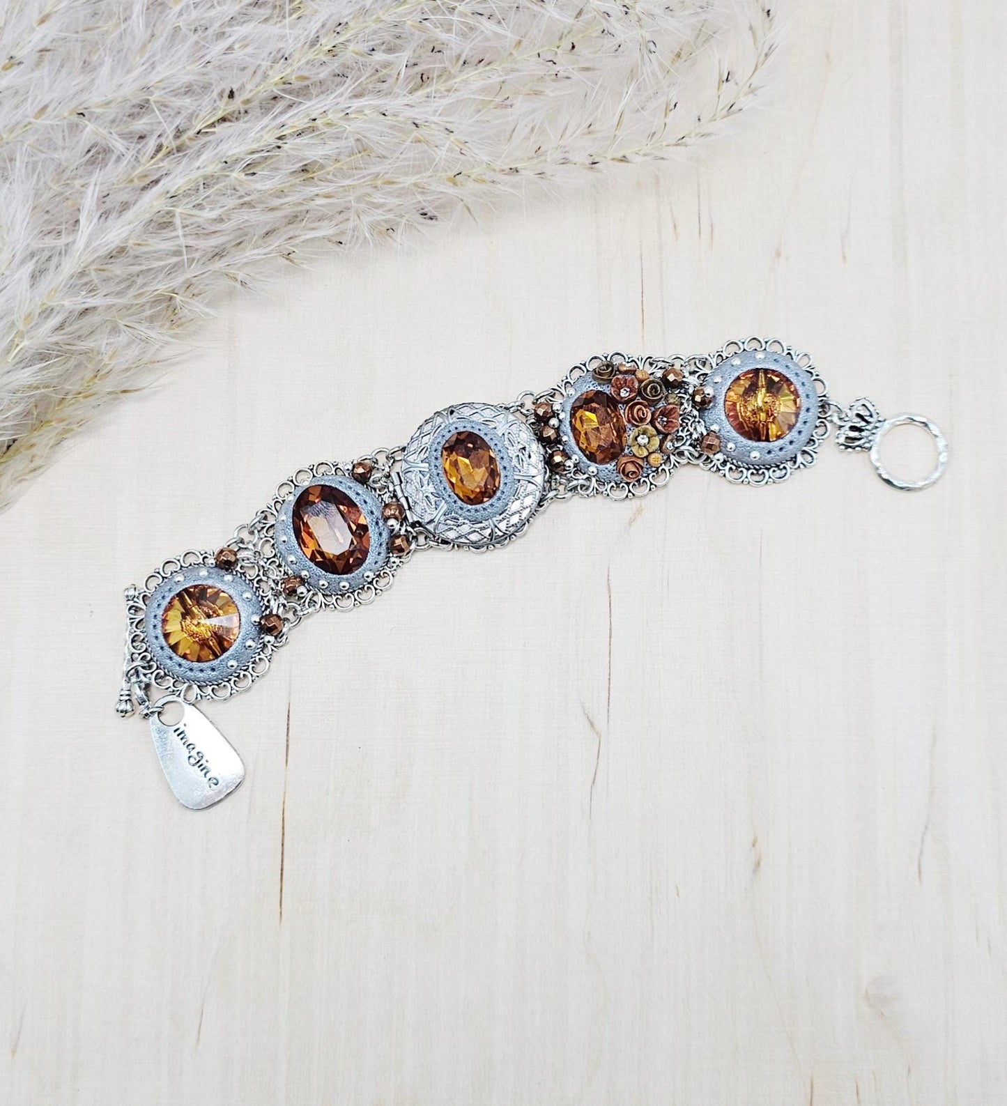 Amber-tone wide bracelet with sparkling crystals and intricate details. Features handmade polymer clay flowers, a functional locket with a red heart inside, and a detachable "imagine" charm.