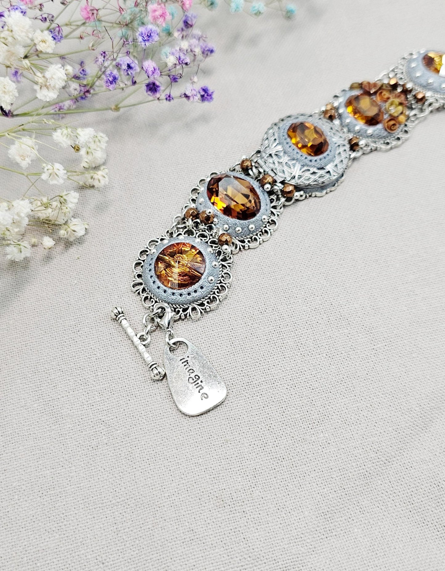 Amber-tone wide bracelet with sparkling crystals and intricate details. Features handmade polymer clay flowers, a functional locket with a red heart inside, and a detachable "imagine" charm.
