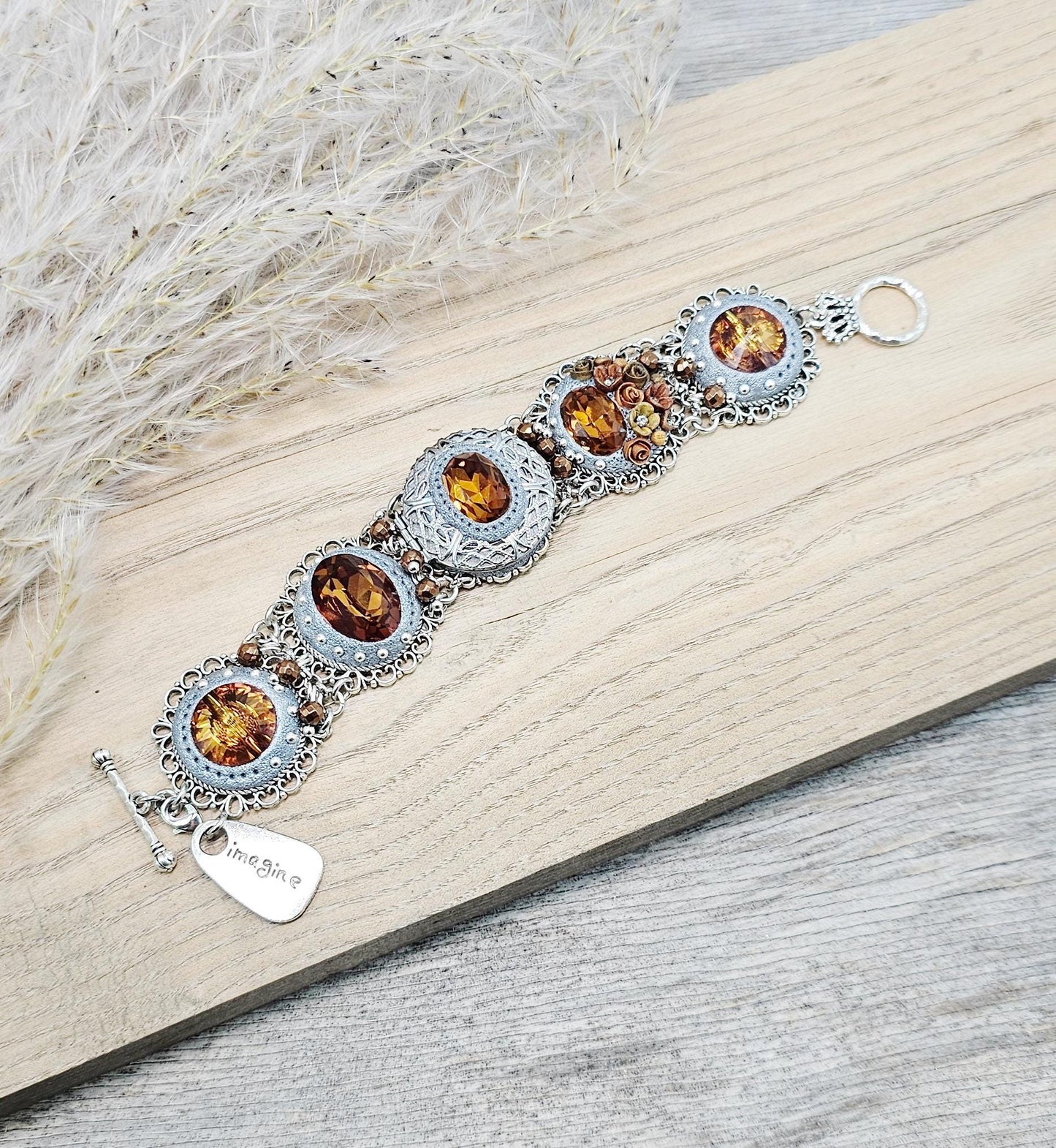 Amber-tone wide bracelet with sparkling crystals and intricate details. Features handmade polymer clay flowers, a functional locket with a red heart inside, and a detachable "imagine" charm.