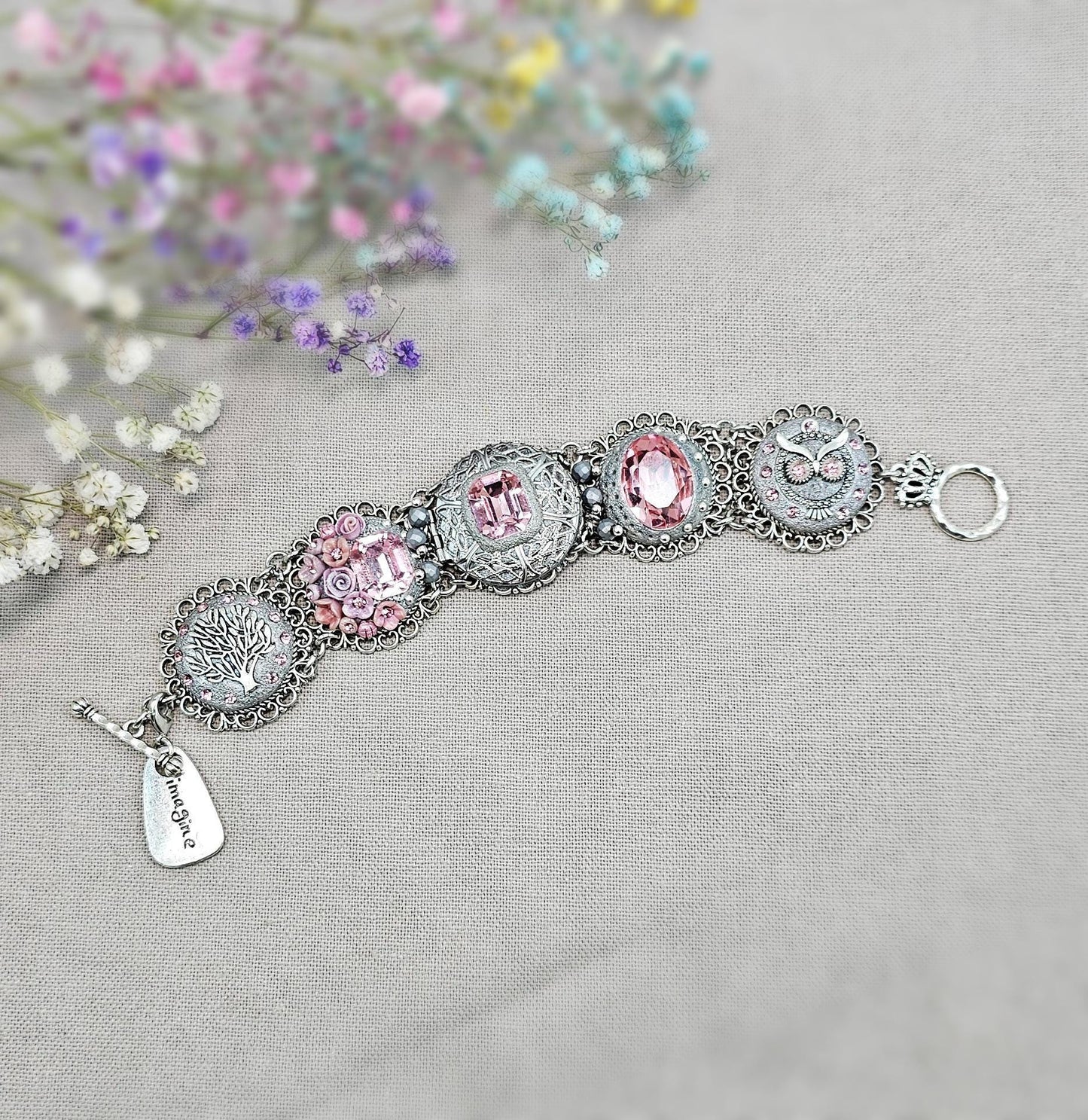 A handmade wide bracelet with pink crystals, polymer clay flowers, a Tree of Life charm, and an owl charm. Features a locket centerpiece with a hidden heart and a removable "Imagine" tag. Elegant and symbolic, perfect as a meaningful gift.