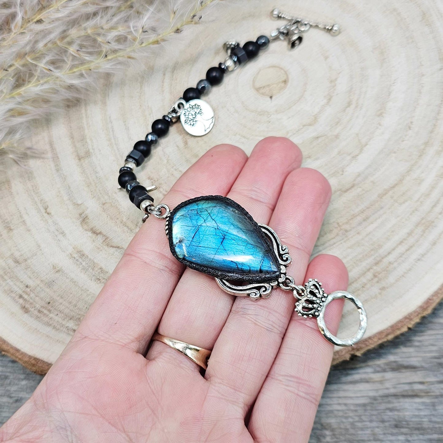 Handmade bracelet featuring a large teardrop labradorite stone with an iridescent blue glow, accented by black hematite beads and silver metal details. Finished with a decorative toggle clasp.