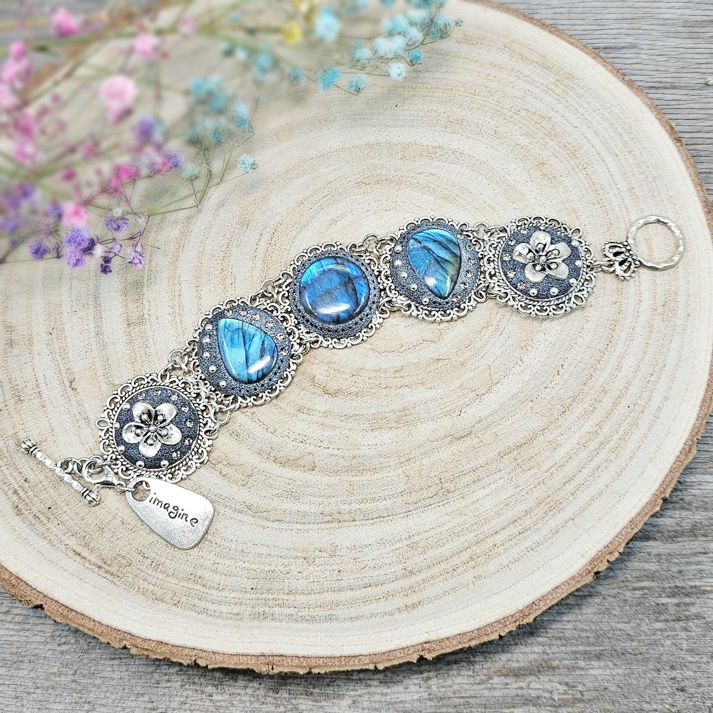 Elegant silver metal bracelet with three shimmering blue labradorite stones and intricate floral details. Features a secure toggle clasp and a detachable "Imagine" charm.