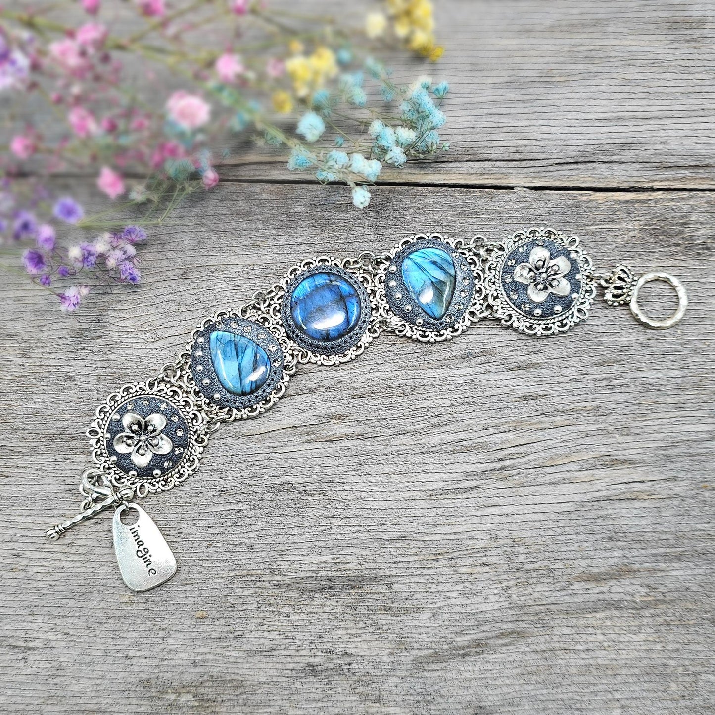 Elegant silver metal bracelet with three shimmering blue labradorite stones and intricate floral details. Features a secure toggle clasp and a detachable "Imagine" charm.