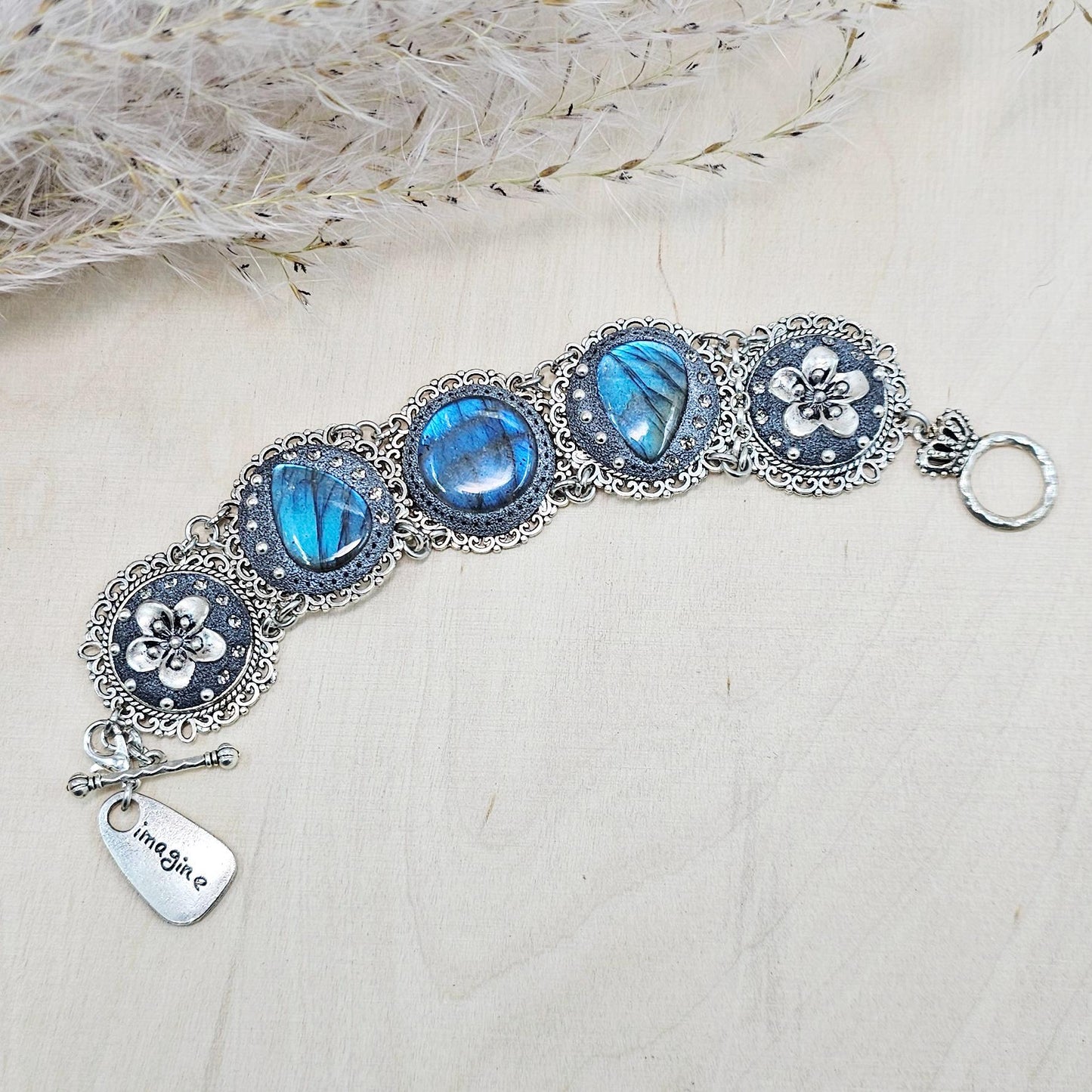 Elegant silver metal bracelet with three shimmering blue labradorite stones and intricate floral details. Features a secure toggle clasp and a detachable "Imagine" charm.