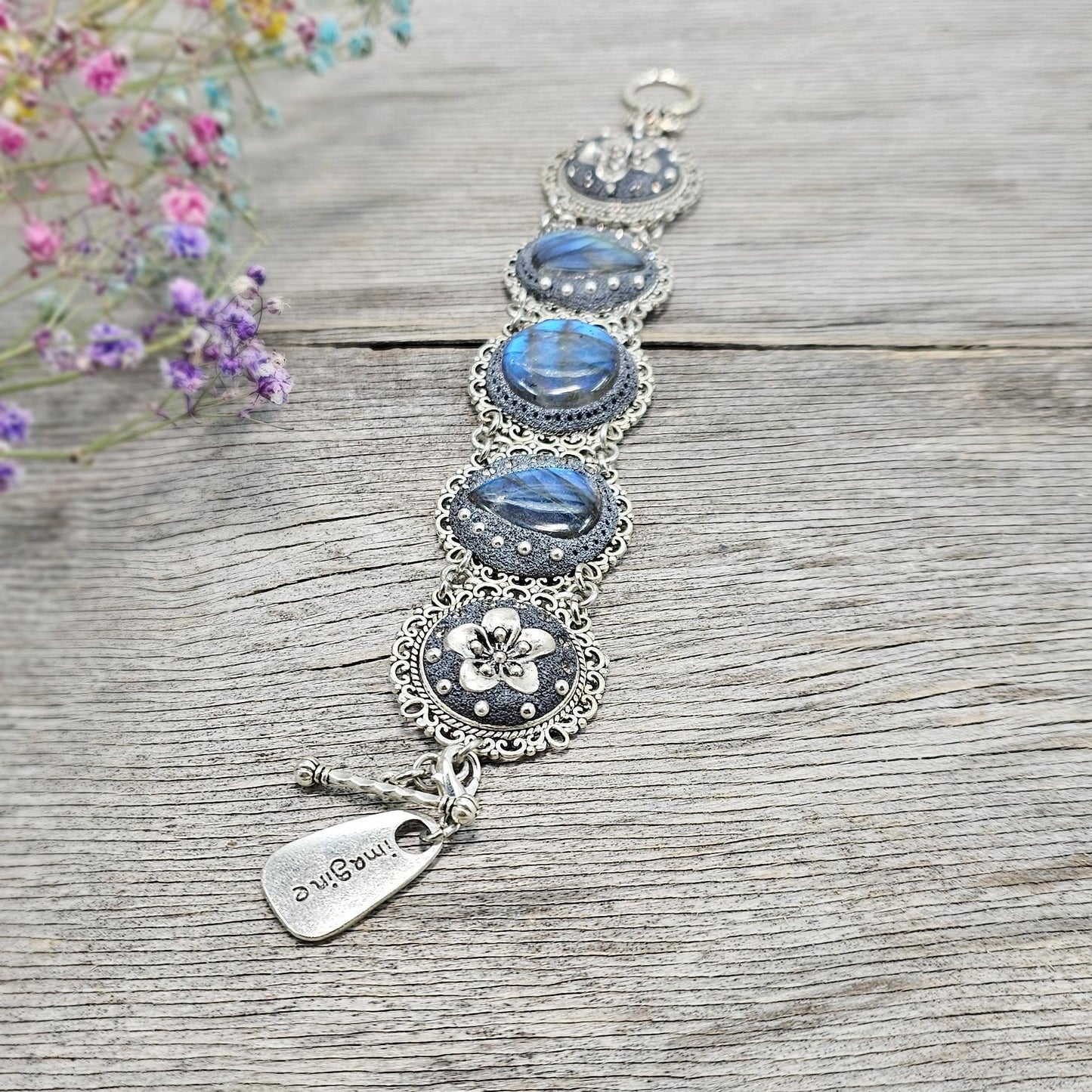 Elegant silver metal bracelet with three shimmering blue labradorite stones and intricate floral details. Features a secure toggle clasp and a detachable "Imagine" charm.