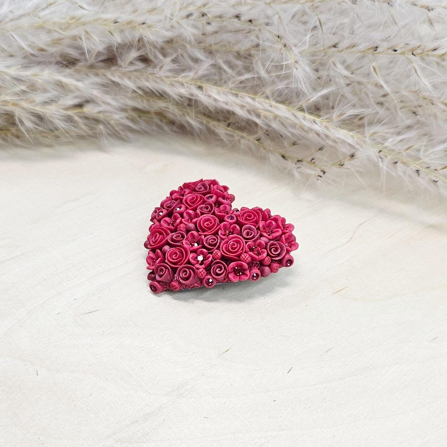 Red heart-shaped brooch made of polymer clay, decorated with hand-sculpted roses and small flowers, accented with crystals.