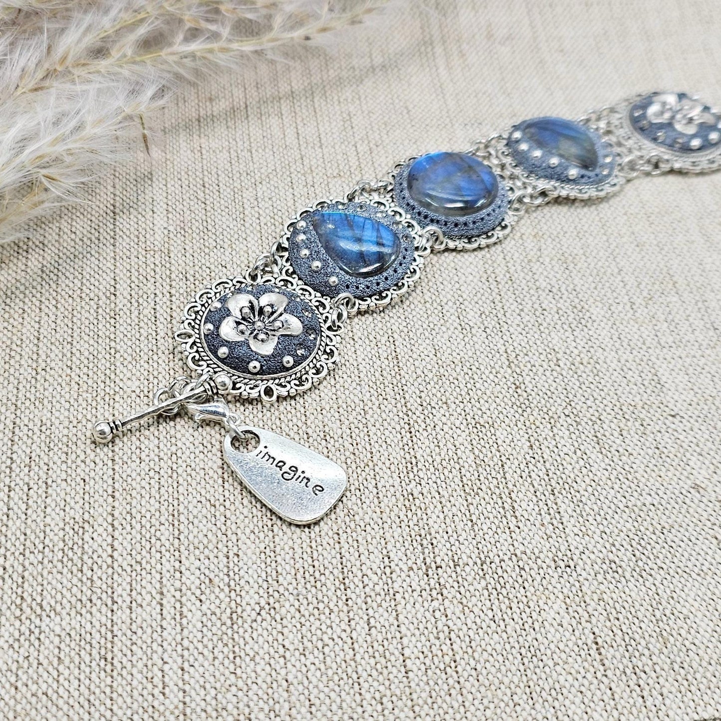 Elegant silver metal bracelet with three shimmering blue labradorite stones and intricate floral details. Features a secure toggle clasp and a detachable "Imagine" charm.