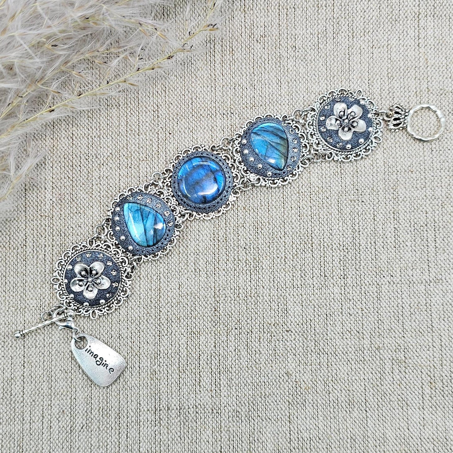 Elegant silver metal bracelet with three shimmering blue labradorite stones and intricate floral details. Features a secure toggle clasp and a detachable "Imagine" charm.