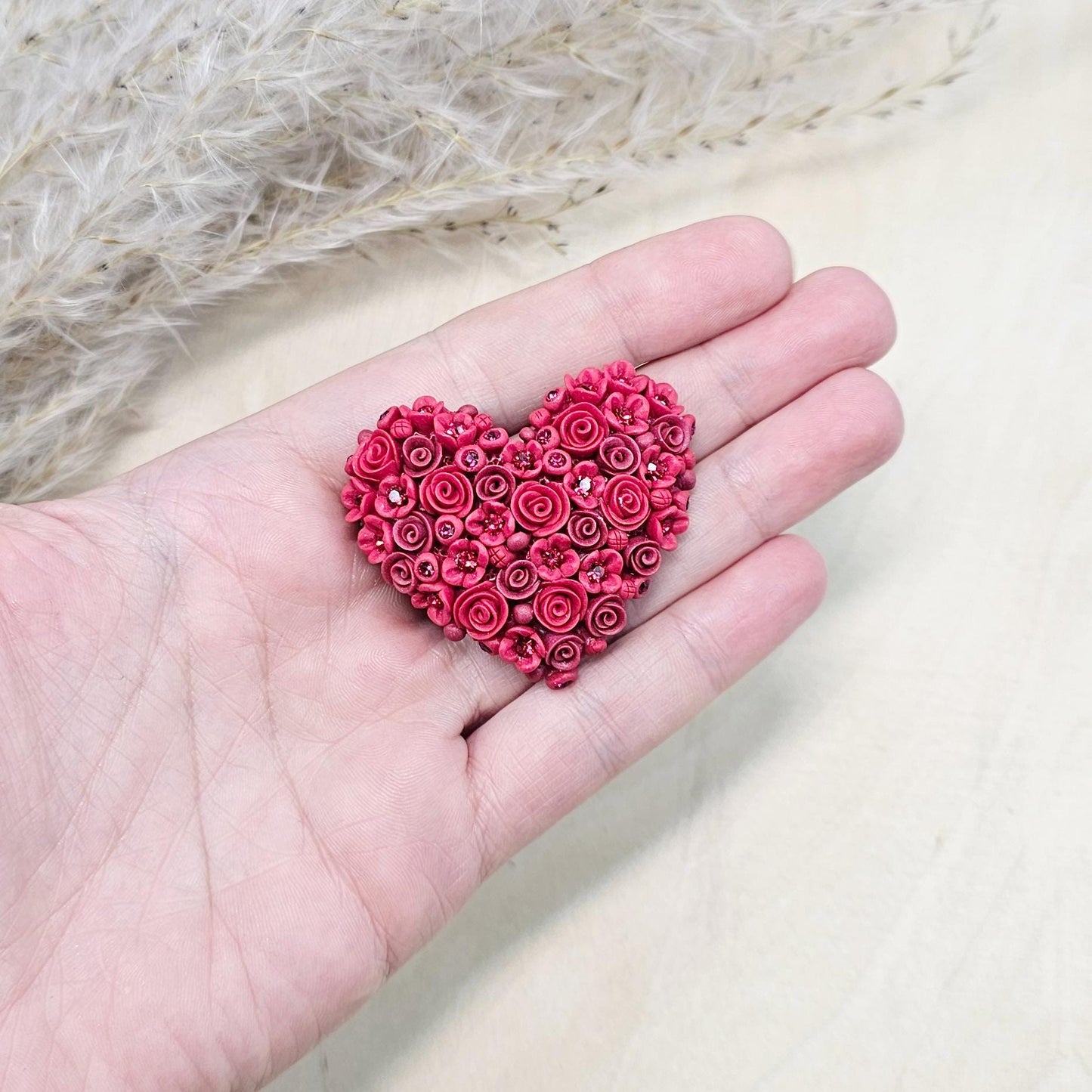 Red heart-shaped brooch made of polymer clay, decorated with hand-sculpted roses and small flowers, accented with crystals.