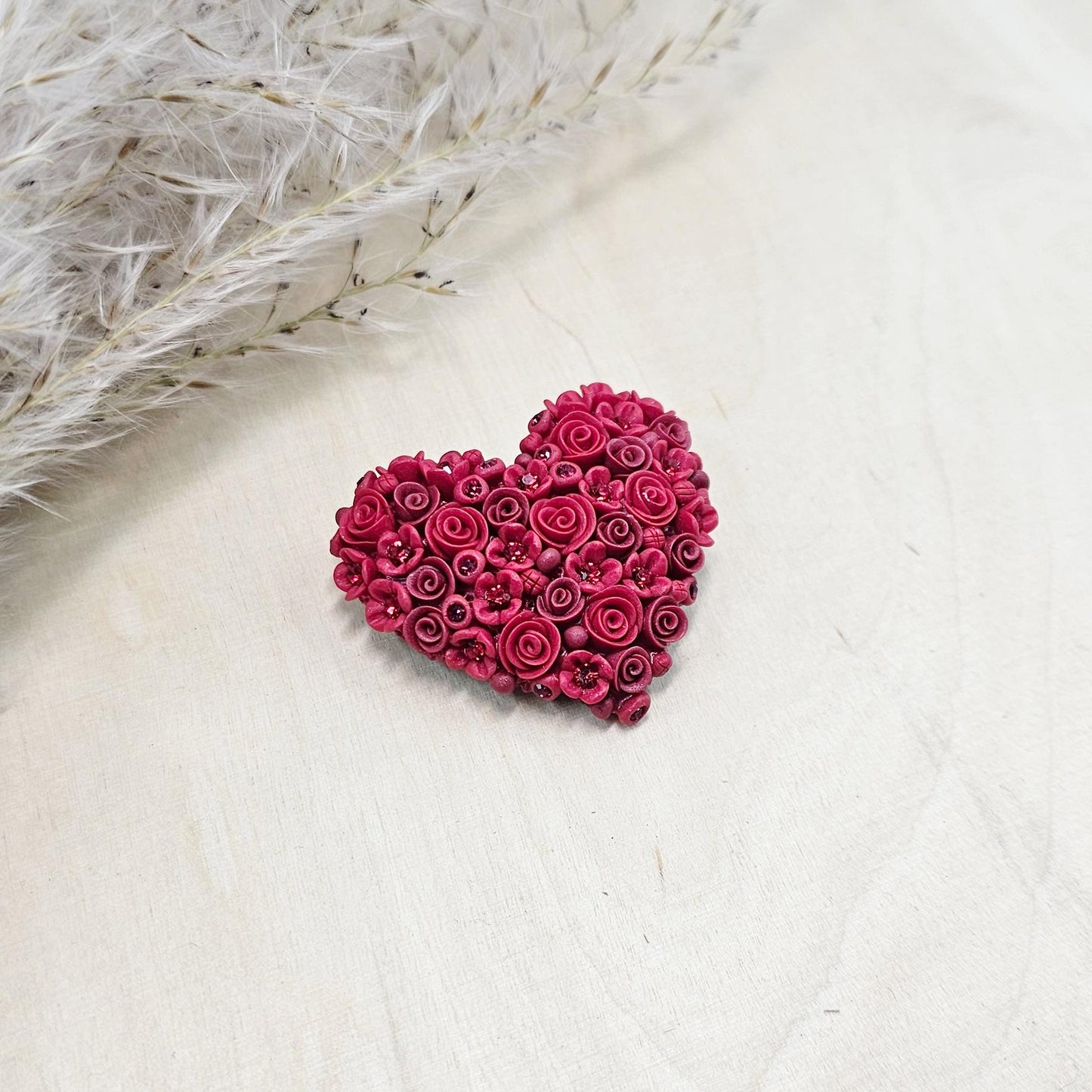 Red heart-shaped brooch made of polymer clay, decorated with hand-sculpted roses and small flowers, accented with crystals.