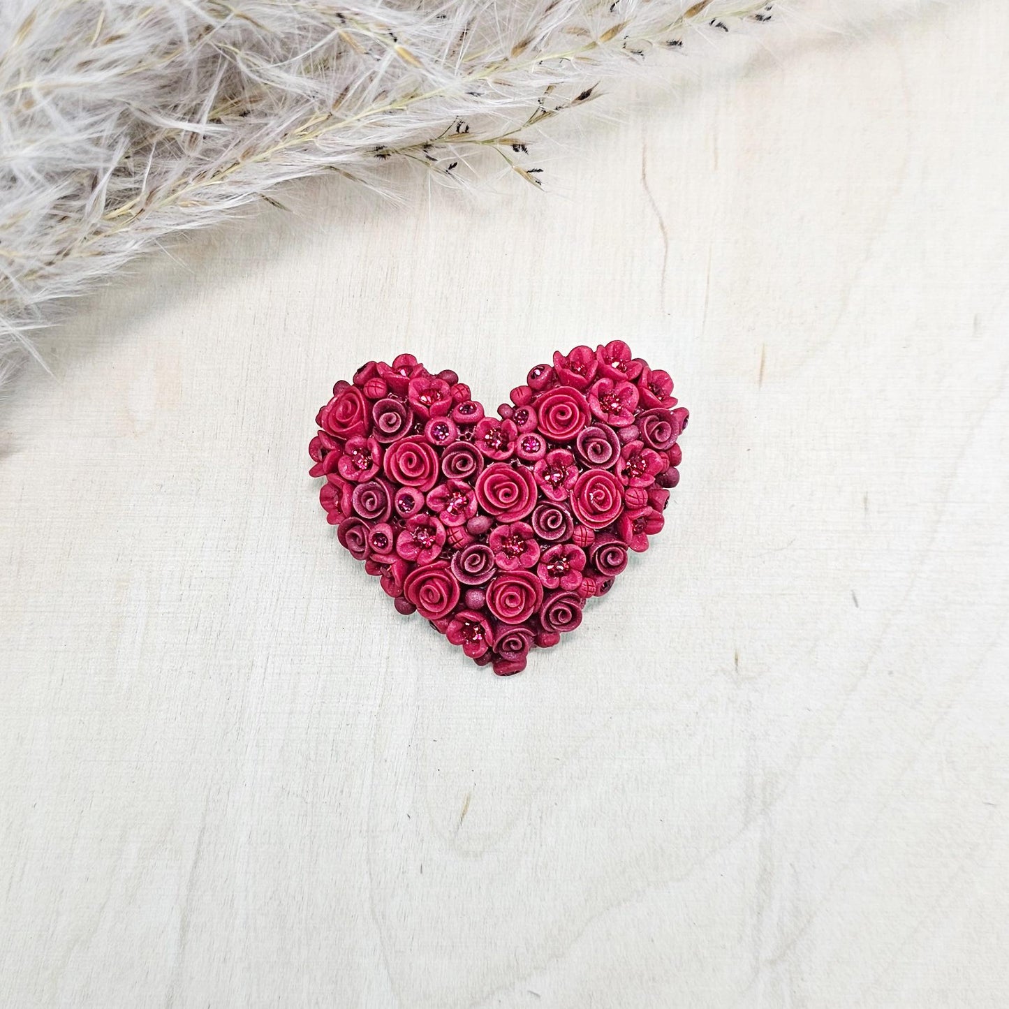 Red heart-shaped brooch made of polymer clay, decorated with hand-sculpted roses and small flowers, accented with crystals.