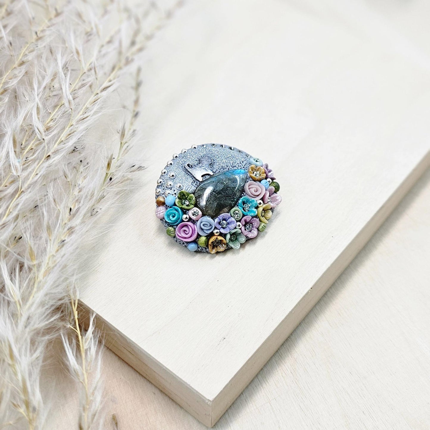 1️⃣ Front view: A handcrafted labradorite brooch featuring a silver-tone cat charm, surrounded by delicate polymer clay flowers in pastel colors. The shimmering gemstone displays a magical glow depending on the lighting.
2️⃣ Back view: The backside of the handmade cat brooch, featuring a secure screw-back pin for comfortable wear. A small red heart detail and subtle embellishments add an extra touch of uniqueness.
Let me know if you need any tweaks! 😊
