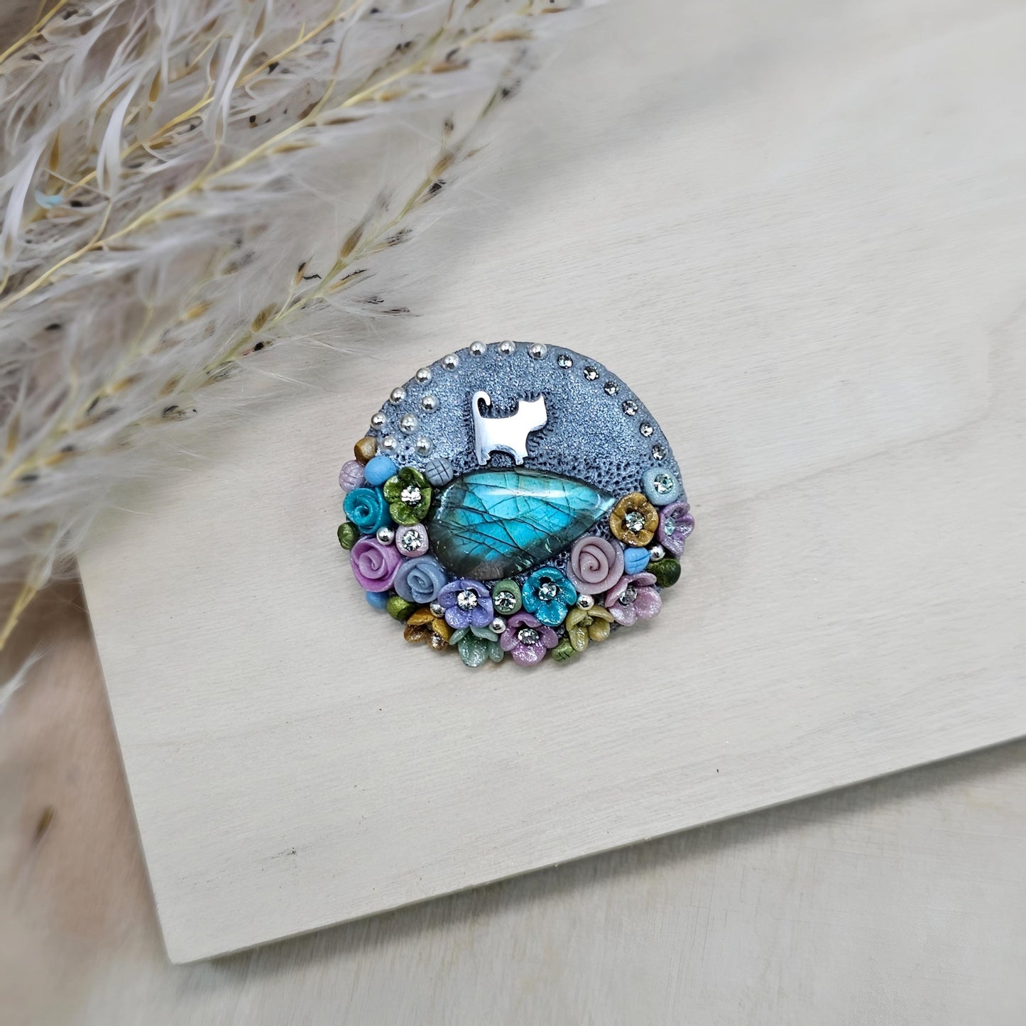 1️⃣ Front view: A handcrafted labradorite brooch featuring a silver-tone cat charm, surrounded by delicate polymer clay flowers in pastel colors. The shimmering gemstone displays a magical glow depending on the lighting.
2️⃣ Back view: The backside of the handmade cat brooch, featuring a secure screw-back pin for comfortable wear. A small red heart detail and subtle embellishments add an extra touch of uniqueness.
Let me know if you need any tweaks! 😊