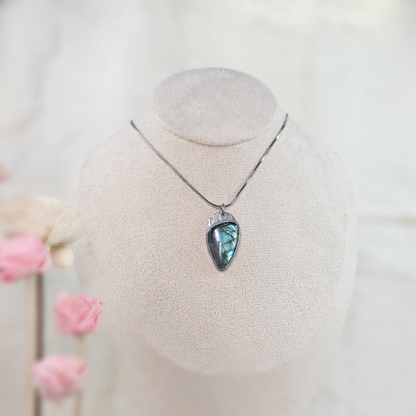 Labradorite pendant displayed on a jewelry bust with soft pink flowers. Elegant handmade necklace for her.